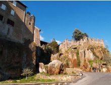 Complesso storico in pietra su uno sperone roccioso, con edifici e una cinta muraria merlata che seguono il profilo della rupe. In primo piano si vede una strada asfaltata in salita con muretto e piccoli spazi verdi, sotto un cielo limpido.