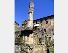 Colonna in pietra di epoca antica, parzialmente danneggiata e rinforzata con anelli metallici, su un basamento in muratura tra rovine e pietrame. Sullo sfondo si vedono edifici in pietra con finestre e persiane in un contesto urbano storico sotto cielo sereno.