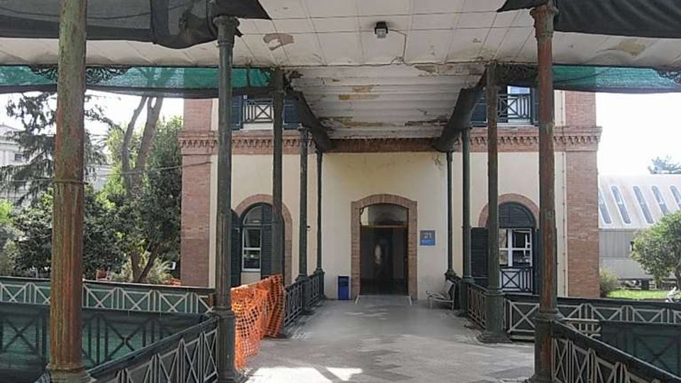Passaggio pedonale coperto con pavimentazione a mosaico, colonne metalliche e ringhiere decorative, che conduce all’ingresso ad arco di un edificio in mattoni e intonaco. La copertura mostra segni di usura e ai lati si intravedono alberi e strutture schermate con teli.