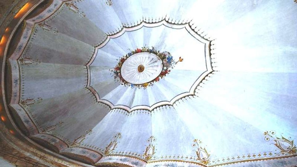 Vista verso l’alto di una cupola interna decorata, con pannelli radiali azzurro chiaro e motivi ornamentali dorati lungo il bordo. Al centro si vede un medaglione ovale con ghirlanda floreale e rosetta, circondato da cornici modanate.