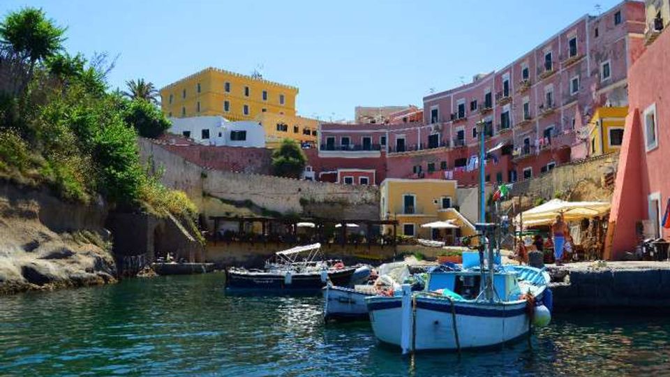 Piccolo porto in una baia rocciosa con acqua calma e diverse barche ormeggiate in primo piano. Sullo sfondo si vedono case a più piani dai colori vivaci su un pendio, un muraglione in pietra e un molo con ombrelloni.