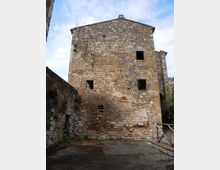 Edificio storico in pietra a tre livelli, con muratura irregolare, crepe visibili e finestre rettangolari prive di infissi. Davanti si vede un piccolo cortile in pietra delimitato da muri laterali, sotto un cielo parzialmente nuvoloso.
