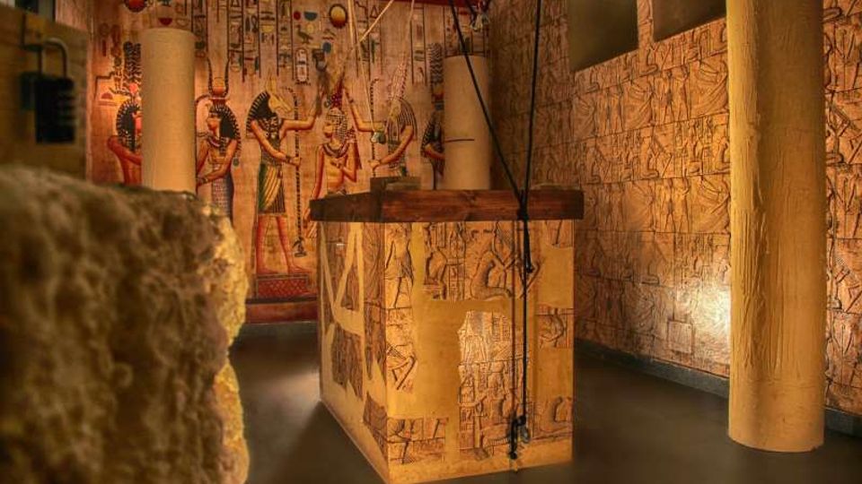 Interno in stile egizio con pareti ricoperte di geroglifici e pitture murali, illuminate da luce calda. Al centro si vede un grande blocco rettangolare decorato e sospeso con cavi, circondato da colonne cilindriche.