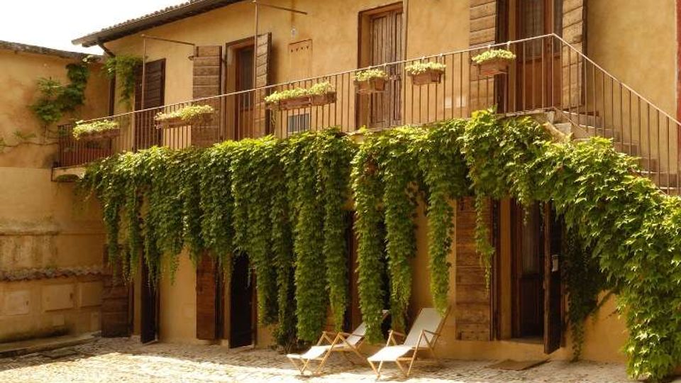 Cortile lastricato in pietra davanti a un edificio rustico a due piani con intonaco beige, balcone con ringhiera metallica e persiane in legno. Tralci di vite pendono dal balcone fino al piano terra e sono presenti sedie a sdraio vicino alle porte.