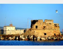 Rovine di un edificio in muratura in riva al mare, con muri in pietra parzialmente crollati e aperture ad arco, circondate da massi frangiflutti. Sullo sfondo si vede una struttura fortificata chiara con torrette e aste per bandiere, sotto un cielo azzurro.