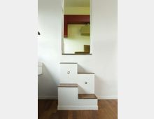 Interno di una casa moderna con piccola scala a gradini sfalsati in muratura bianca e pedate in legno, illuminata da luci incassate. In alto si apre un grande vano rettangolare che affaccia su un soppalco con elementi d’arredo e pareti chiare.