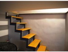 Scala interna compatta con gradini in legno a sbalzo, fissati a una struttura metallica contro una parete grigia. L’ambiente ha soffitto basso e illuminazione dall’alto, con pavimento in piastrelle scure.