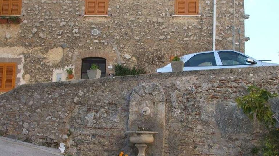 Fontana in pietra con vasca su gradini semicircolari lungo una strada in pendenza, addossata a un muro di contenimento in muratura. Sullo sfondo si vede una casa in pietra a due piani con persiane in legno e tetto in coppi.