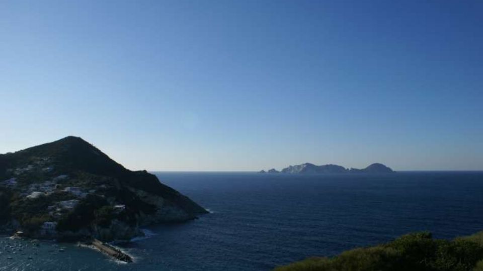 Panorama costiero con un promontorio roccioso e vegetazione in primo piano, una piccola baia con costruzioni ai piedi della collina e il mare aperto. All’orizzonte si vedono alcune isole basse sotto un cielo sereno.