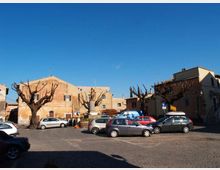 Piazza urbana lastricata con parcheggio di auto, circondata da edifici in muratura dai toni ocra e da alberi potati senza foglie. Al centro si vede una piccola aiuola con un monumento, sotto un cielo sereno.