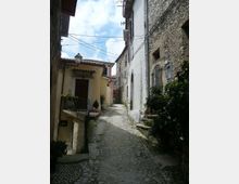 Vicolo stretto in pietra con pavimentazione acciottolata che sale tra case in muratura e intonaco, con finestre ad arco e piccoli balconi. Lampione a parete, cavi sospesi e vasi di piante lungo i muri sotto un cielo parzialmente nuvoloso.