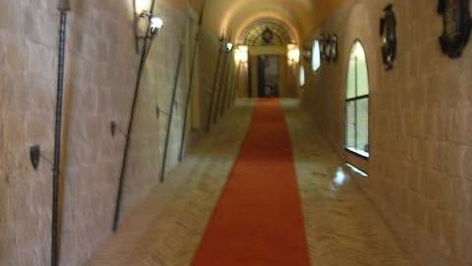Lungo corridoio in pietra con volta a botte e pavimento in mattoni, attraversato da un tappeto rosso centrale che conduce a una porta in fondo. Sulle pareti sono appese armi decorative e lampade, con alcune finestre ad arco sul lato destro.