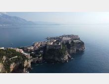Veduta aerea di un promontorio roccioso sul mare con una città costiera compatta e una grande fortezza in pietra affacciata su acque calme. Sullo sfondo si intravedono montagne e una linea di costa lontana.