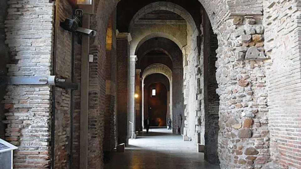 Corridoio interno in muratura antica con una sequenza di grandi archi in mattoni e pietra, pavimento in lastre di pietra e luci a parete. La prospettiva conduce verso aperture ad arco successive, creando un passaggio lungo e stretto in stile storico.