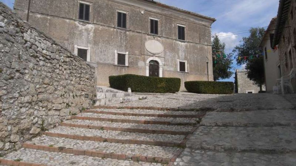 Scalinata in pietra e ciottoli che conduce a un edificio storico in intonaco grigio, con porta centrale ad arco, cornici semplici e finestre rettangolari. Ai lati si vedono un muretto in pietra a secco, siepi basse potate e una stretta strada in salita tra edifici.