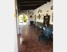 Veranda coperta in stile rustico con pavimento in cotto, soffitto con travi in legno scuro e pareti bianche decorate con piatti e cornici. Lungo la parete ci sono tavoli e sedie in plastica e un mobile credenza; un grande arco aperto conduce al giardino esterno.
