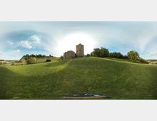 Panorama a 360° di una collina erbosa con un antico edificio in pietra tipo castello o monastero, con torre centrale e strutture annesse. Intorno si vedono campi ondulati e gruppi di alberi sotto un cielo parzialmente nuvoloso.