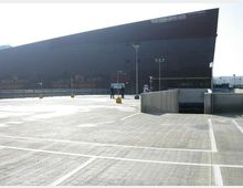 Ampia area pavimentata tipo parcheggio o piazzale, con segnaletica a linee bianche, lampioni e una rampa di accesso in cemento. Sullo sfondo si vede un grande edificio moderno scuro con facciata inclinata e superfici lisce.