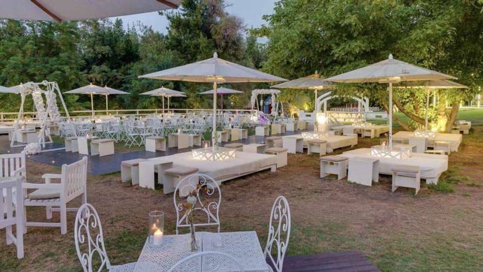 Area lounge all’aperto in un giardino con divanetti e tavolini bianchi, ombrelloni grandi e sedute in ferro battuto. Sullo sfondo si vedono una pedana con molte sedie disposte in file e alberi fitti che circondano lo spazio.