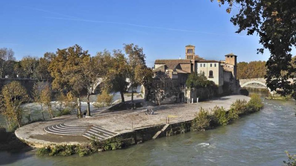 Isola fluviale con una piazza pavimentata e gradinate semicircolari affacciate sull’acqua, circondata da alberi. Sullo sfondo si vedono edifici storici con una torre e un ponte ad arco che attraversa il fiume.