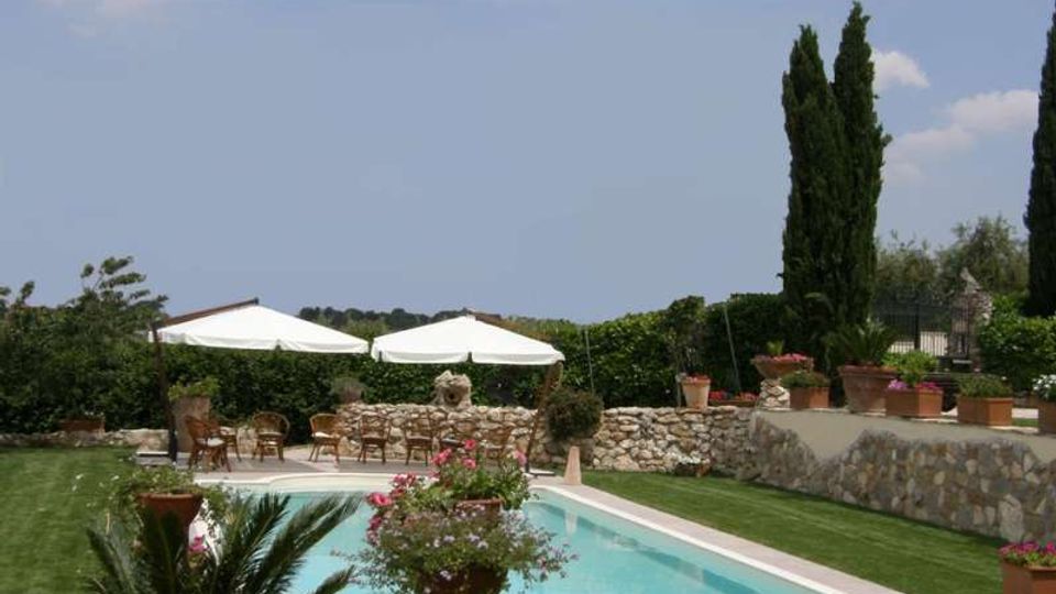 Piscina rettangolare all’aperto con bordo chiaro, circondata da prato curato e muretti in pietra con vasi di fiori. Sullo sfondo ci sono due grandi ombrelloni bianchi con sedie, siepi e alti cipressi sotto un cielo parzialmente nuvoloso.
