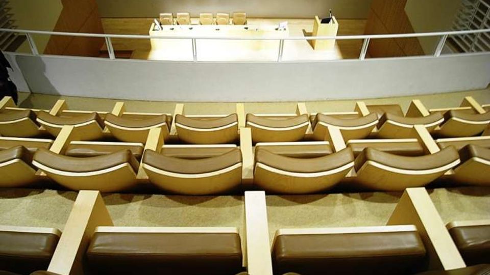 Interno di un’aula magna o auditorium con file di sedute imbottite color beige disposte in gradoni e rivolte verso un palco. In fondo si vede un lungo tavolo con sedie e un leggio, separati da una balaustra chiara.