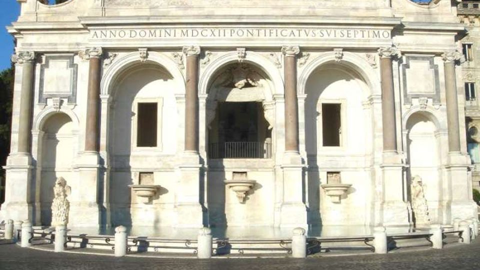 Facciata monumentale in pietra chiara di una fontana storica, con tre grandi arcate e colonne, iscrizioni in latino nella parte superiore e vasche frontali. Davanti si vede una piazza pavimentata in sampietrini con paletti di delimitazione.