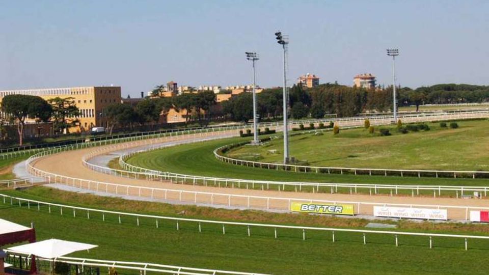 Ippodromo all’aperto con pista ovale in sabbia e prato, delimitata da ringhiere bianche e illuminata da alti fari. Sullo sfondo si vedono edifici bassi e alberi sotto un cielo sereno.