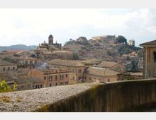 Veduta panoramica di un borgo collinare con case dai tetti in coppi e edifici storici addossati, tra cui una torre campanaria e una chiesa. In primo piano si vede un muretto in pietra, con il centro abitato che si estende sulle pendici sotto un cielo parzialmente nuvoloso.