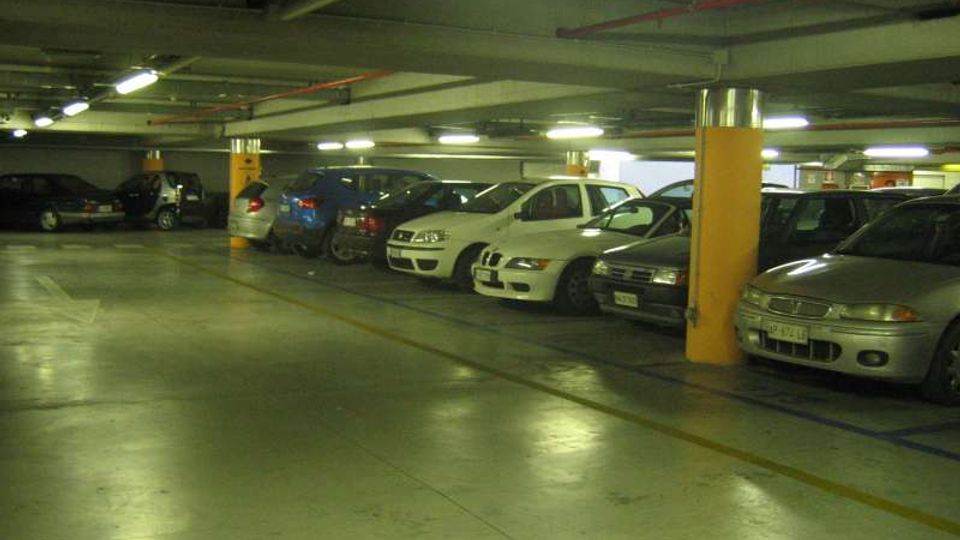 Parcheggio sotterraneo con file di auto parcheggiate lungo le pareti e una corsia centrale libera. Soffitto basso con tubazioni a vista e illuminazione al neon, pilastri con fasce gialle e pavimento in cemento con strisce di delimitazione.