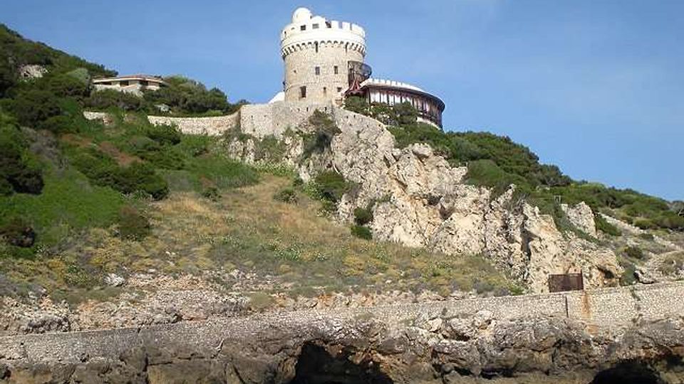 Castello in pietra con torre cilindrica e merli, arroccato su una scogliera rocciosa sopra il mare. Il pendio è coperto di vegetazione mediterranea e muri di contenimento, con un edificio adiacente e cielo sereno sullo sfondo.