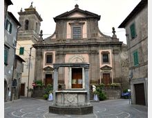 Facciata di una chiesa storica in pietra e intonaco rosa, con portale centrale incorniciato, due ingressi laterali e campanile sul lato sinistro, affacciata su una piccola piazza. In primo piano si vede una fontana ottagonale in pietra al centro della pavimentazione circolare, circondata da edifici residenziali.