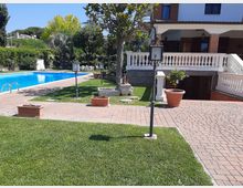 Giardino di una villa con piscina rettangolare azzurra, vialetti in mattoni e prato curato con lampioni e vasi. Sullo sfondo si vede la casa con veranda coperta, ringhiera bianca e scale, circondata da alberi e siepi.