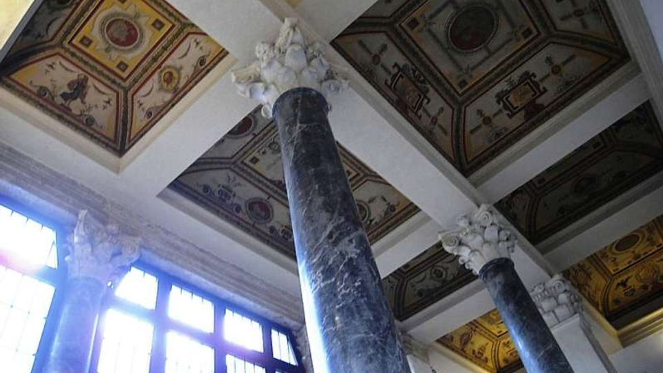 Interno di un edificio storico con alte colonne in marmo scuro e capitelli decorati, affacciate su grandi finestre che lasciano entrare molta luce. Il soffitto a cassettoni è suddiviso da travi bianche e presenta riquadri affrescati con motivi ornamentali.