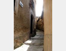 Vicolo stretto in un centro storico, con pavimentazione in pietra e alcuni gradini, fiancheggiato da alte pareti intonacate. Si vedono un portale ad arco, un piccolo balcone in ferro battuto e un lampione lungo il passaggio.