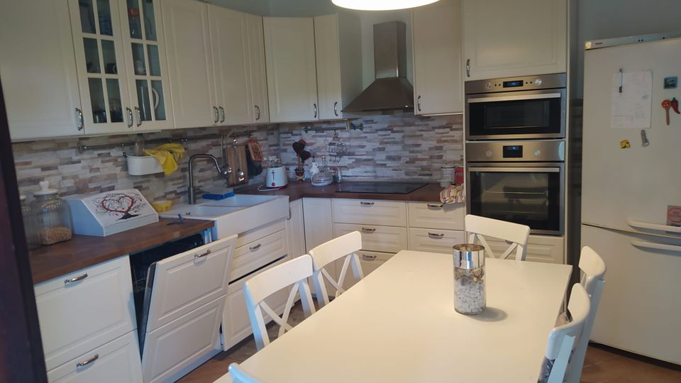 Cucina domestica luminosa con mobili bianchi a parete, piano di lavoro in legno e paraschizzi a mosaico in tonalità beige e grigie. In primo piano c’è un tavolo da pranzo bianco con sedie, mentre sul lato destro si vedono forno e microonde incassati e un grande frigorifero.