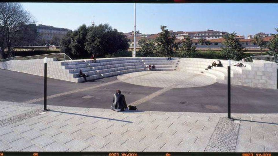 Anfiteatro all’aperto in pietra chiara con gradoni semicircolari e area centrale pavimentata, inserito in una piazza con lampioni e vialetti. Sullo sfondo si vedono alberi e edifici urbani sotto un cielo sereno.