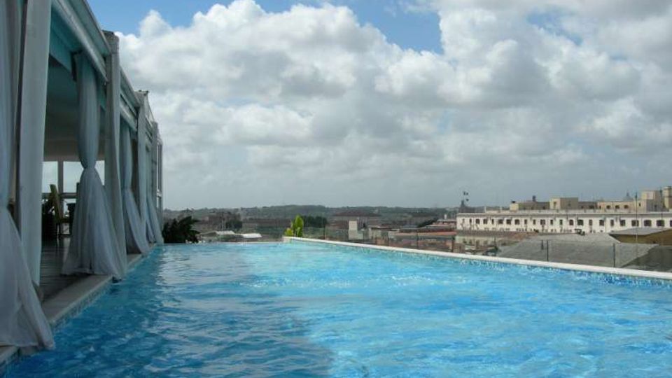 Piscina a sfioro su una terrazza panoramica, affiancata da una struttura con vetrate e tende bianche. Sullo sfondo si vedono tetti e edifici della città sotto un cielo parzialmente nuvoloso.