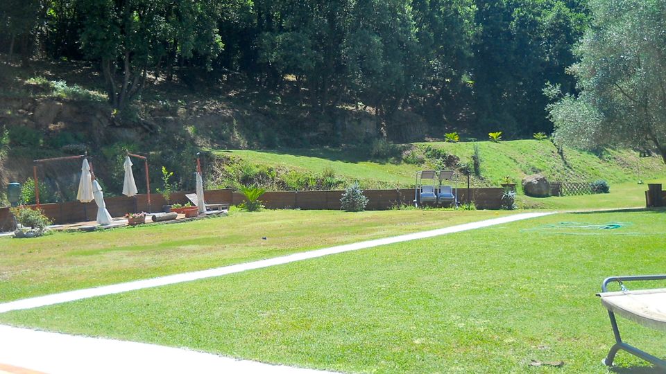 Ampio prato verde in un’area all’aperto con vialetto chiaro curvo, aiuole e ombrelloni chiusi vicino a una piccola zona pavimentata con sedie. Sullo sfondo c’è un terrapieno e una collina boscosa con alberi fitti e vegetazione.