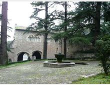 Cortile lastricato con una fontana centrale e alti alberi sempreverdi, affacciato su un edificio storico in pietra con porticati ad archi e finestre ad arco. Sullo sfondo si vedono muri e un parapetto in pietra che delimitano lo spazio esterno.