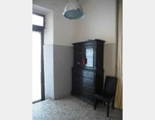 Interno di una stanza spoglia con pareti bianche e grigie e pavimento in graniglia, illuminata da una porta finestra sulla sinistra. Nell’angolo c’è una credenza scura in legno e, accanto, una sedia nera; dal soffitto pende una lampada a sospensione.