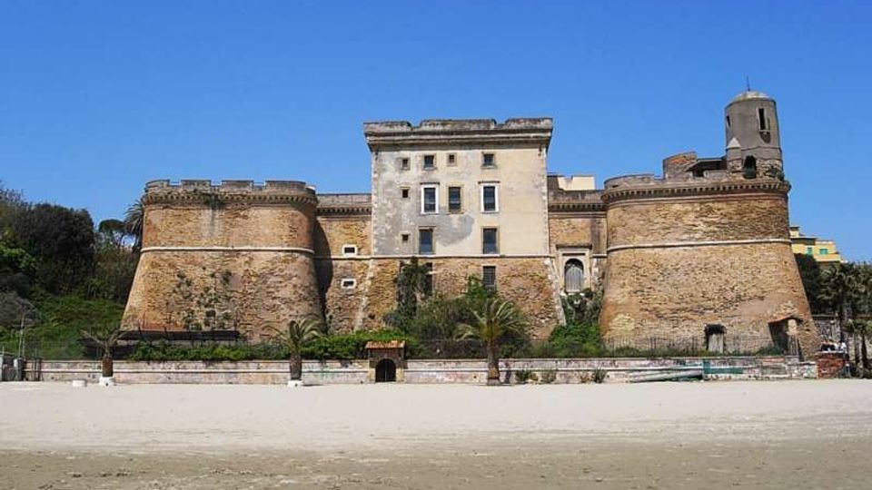 Castello costiero in pietra con due grandi bastioni cilindrici e corpo centrale rettangolare con finestre, affacciato su una spiaggia sabbiosa. Sullo sfondo si vede cielo sereno e vegetazione con palme lungo il margine della fortificazione.