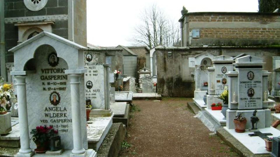 Viale centrale di un cimitero con tombe in marmo e cappelle funerarie disposte ai lati, con vasi di fiori e lastre commemorative. Sullo sfondo si vedono un edificio in muratura con facciata decorata e un alto muro perimetrale con alberi.