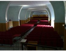 Interno di una sala auditorium con soffitto a volta e grandi archi laterali, pareti chiare e rivestimento in legno lungo i lati. File di poltroncine rosse disposte in platea con corridoio centrale e palco rialzato sul fondo.
