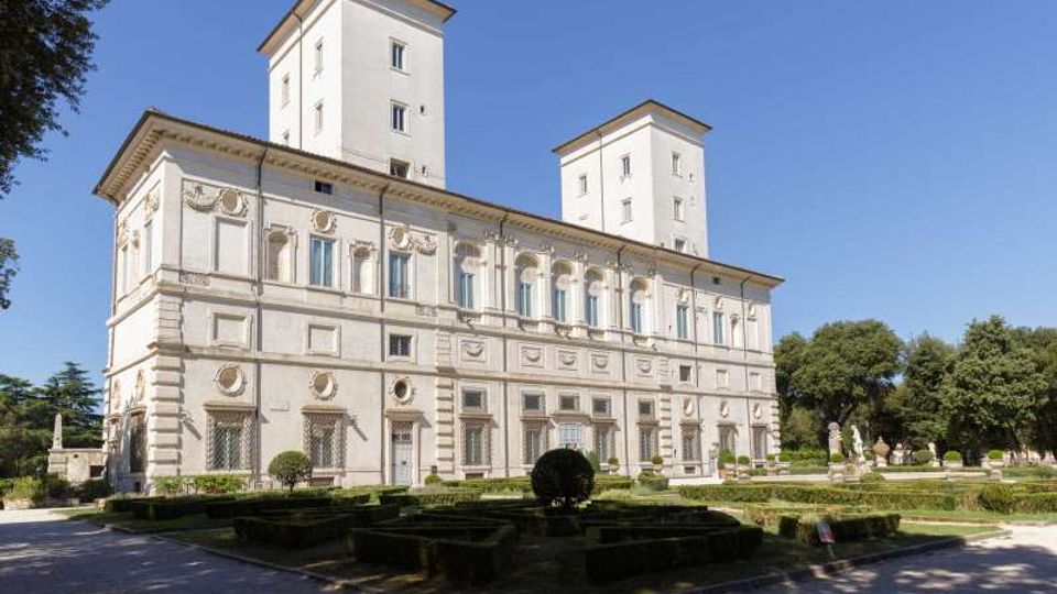 Grande palazzo storico in stile rinascimentale con facciata chiara decorata da cornici e finestre regolari, affiancato da due torri rettangolari. In primo piano si vede un giardino geometrico con siepi basse e vialetti, circondato da alberi sotto un cielo sereno.