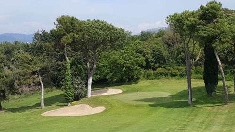 Campo da golf con fairway in erba rasata, green e due bunker di sabbia, circondato da alberi alti e vegetazione fitta. Sullo sfondo si intravedono colline sotto un cielo sereno.