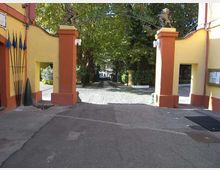 Ingresso con cancello aperto tra due pilastri intonacati giallo e rosso, sormontati da statue e collegati da un arco in ferro. Oltre il varco si vede un viale lastricato che conduce in un’area alberata.