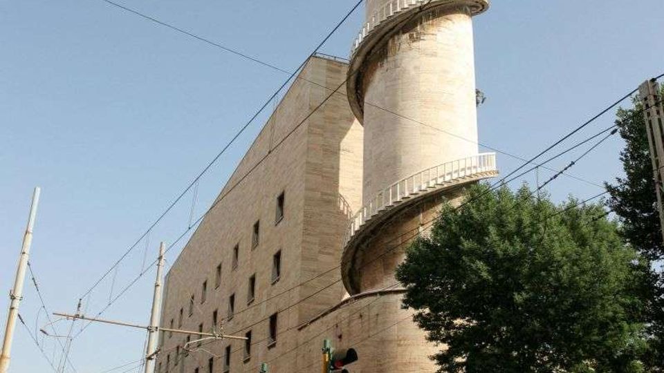 Edificio industriale in muratura chiara affacciato su una strada urbana con binari del tram e cavi aerei, dominato da un’alta torre cilindrica con scala a spirale esterna e ringhiere. In primo piano si vedono semafori e un albero lungo il marciapiede.