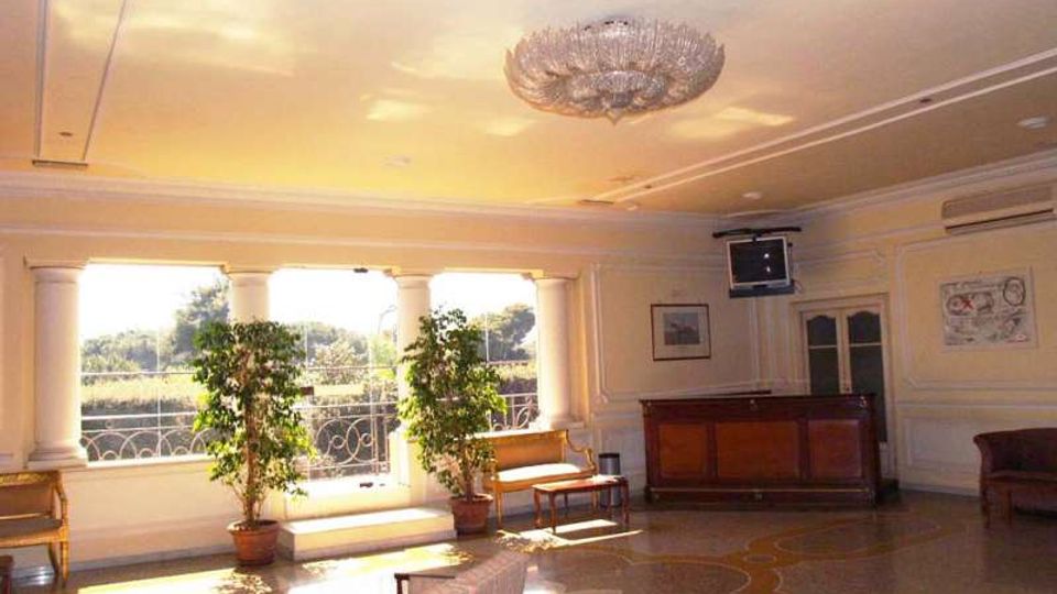 Ampia sala interna in stile classico con grandi finestre ad arco e ringhiera decorativa, pavimento a mosaico e lampadario centrale. Sono presenti panche e divani lungo le pareti, piante in vaso vicino alle finestre e un bancone in legno in un angolo.