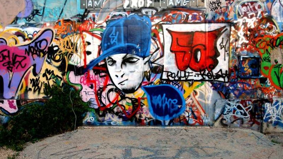Muro esterno in cemento ricoperto da graffiti e murales multicolori, con un grande disegno centrale di un volto stilizzato con cappellino blu e numerose scritte sovrapposte. Davanti al muro c’è un’area pavimentata in cemento con segni e vernice, ai margini qualche ciuffo di vegetazione.
